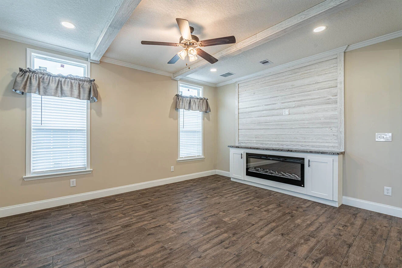A bright empty room with two tall windows, wood-look flooring, a ceiling fan with lights, and a built-in cabinet and electric fireplace on one wall.