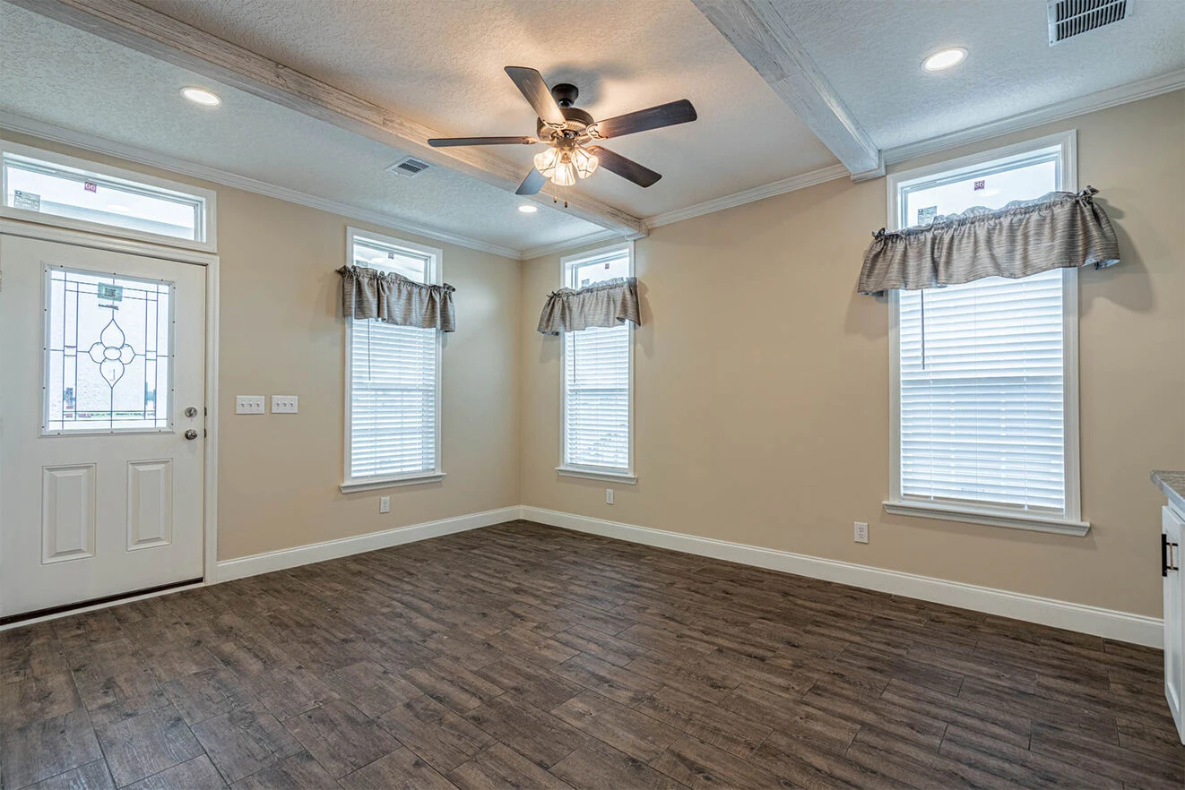 Empty living room with wood-style flooring, three tall windows with valances, a ceiling fan with lights, and a white front door with decorative glass.