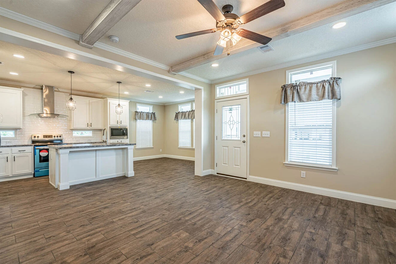 Open-concept living area with wood-look flooring, ceiling beams, ceiling fan, large windows, and a modern kitchen featuring white cabinets and pendant lighting.