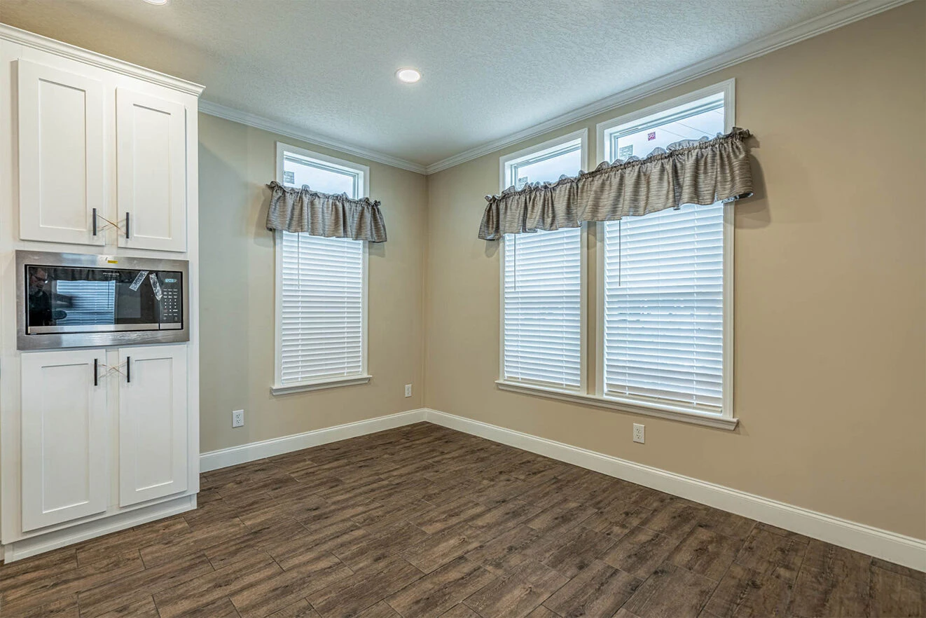 Empty dining area with three tall windows, white blinds, gray valances, beige walls, wood flooring, and a corner of white kitchen cabinets with a built-in microwave.
