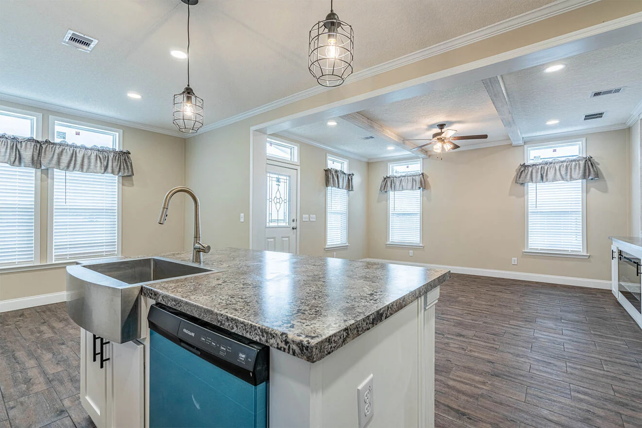 Modern open-concept kitchen and living area with granite island, farmhouse sink, blue dishwasher, ceiling lights, and large windows with valances.