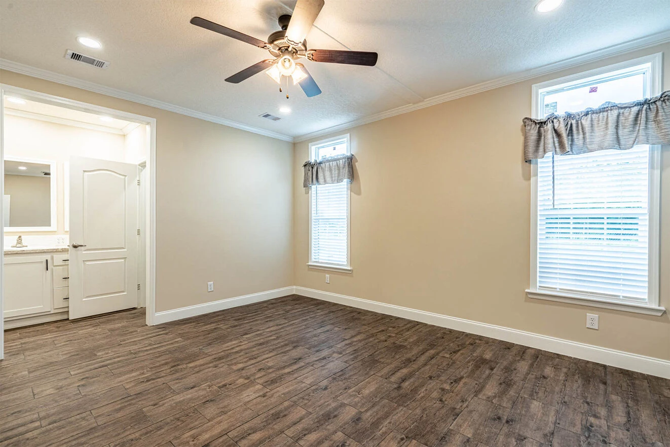 Empty room with wood-look flooring, beige walls, ceiling fan with lights, two windows with curtains, and an open doorway leading to a bathroom.