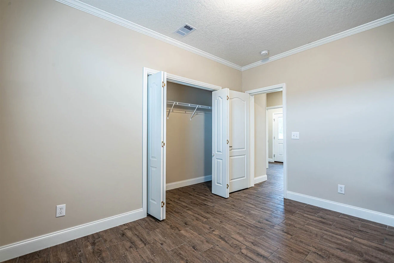Empty room with light beige walls, a double-door closet, wood-style flooring, and white trim. The closet has a single shelf and hanging rod. An open doorway leads to a hallway.