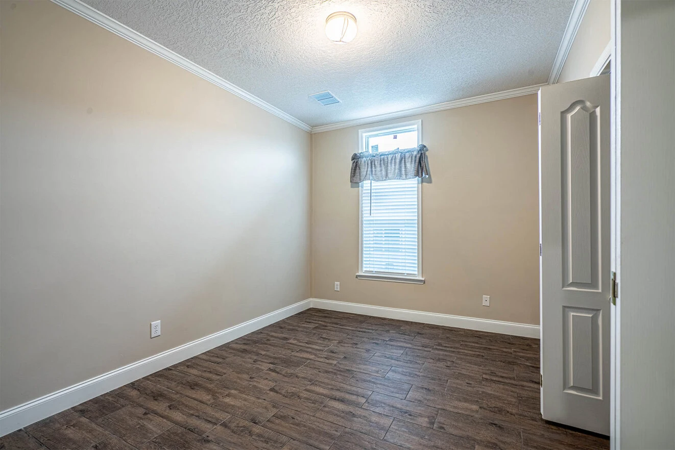 Empty room with beige walls, white trim, a small window with a valance, wood-look flooring, and an open white door. Ceiling light is on.