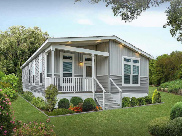 A modern manufactured home with a small front porch, steps leading up, landscaped yard, and trees in the background.