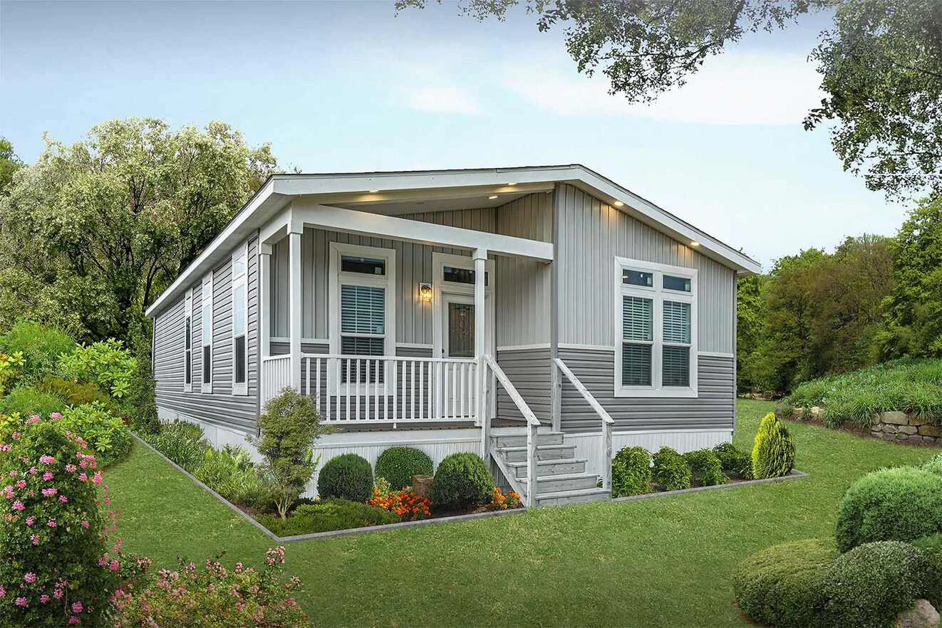 A modern manufactured home with a small front porch, steps leading up, landscaped yard, and trees in the background.