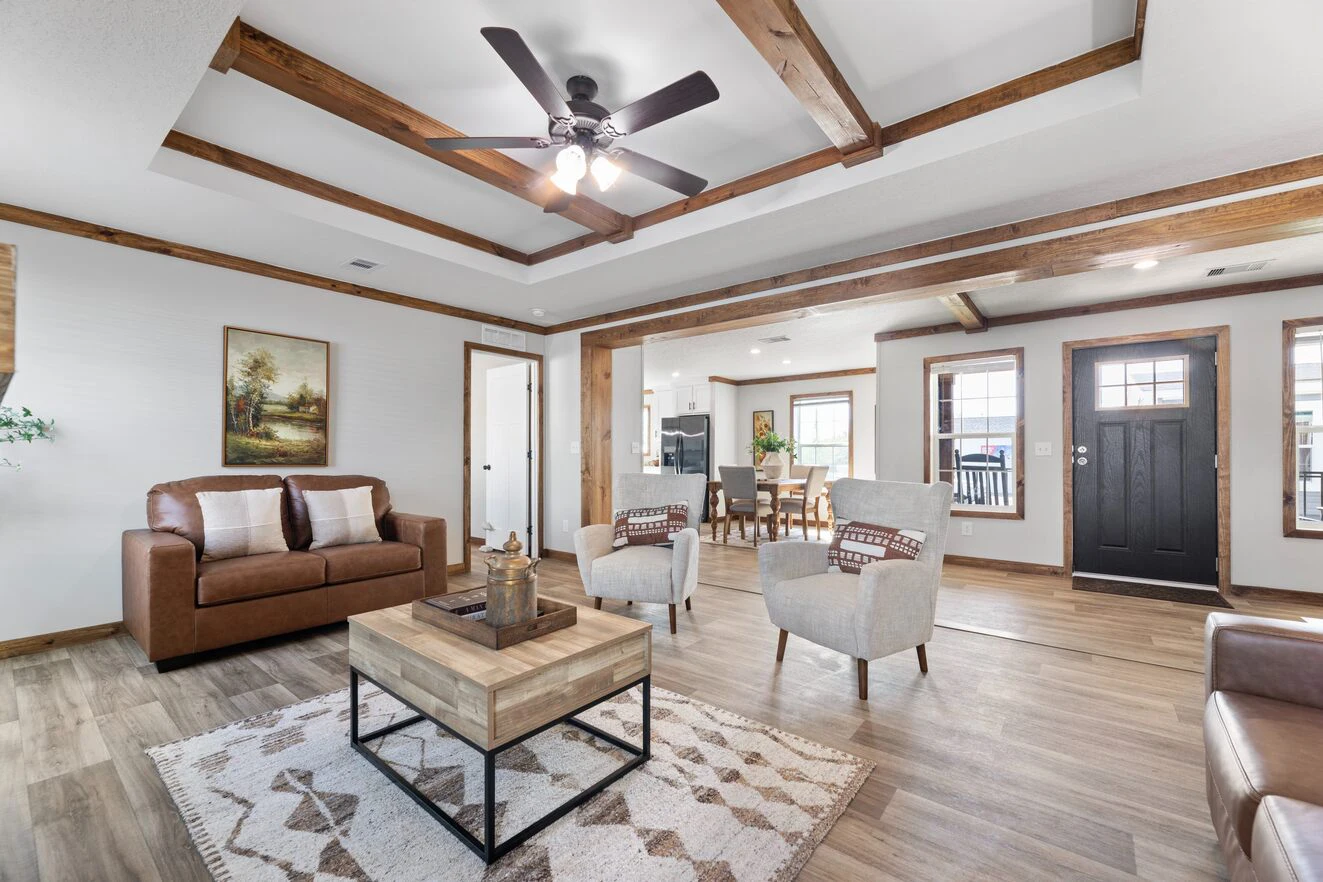 Spacious living room with wooden beams, ceiling fan, brown sofa, two gray armchairs, a coffee table, rug, and view into dining area and front door.