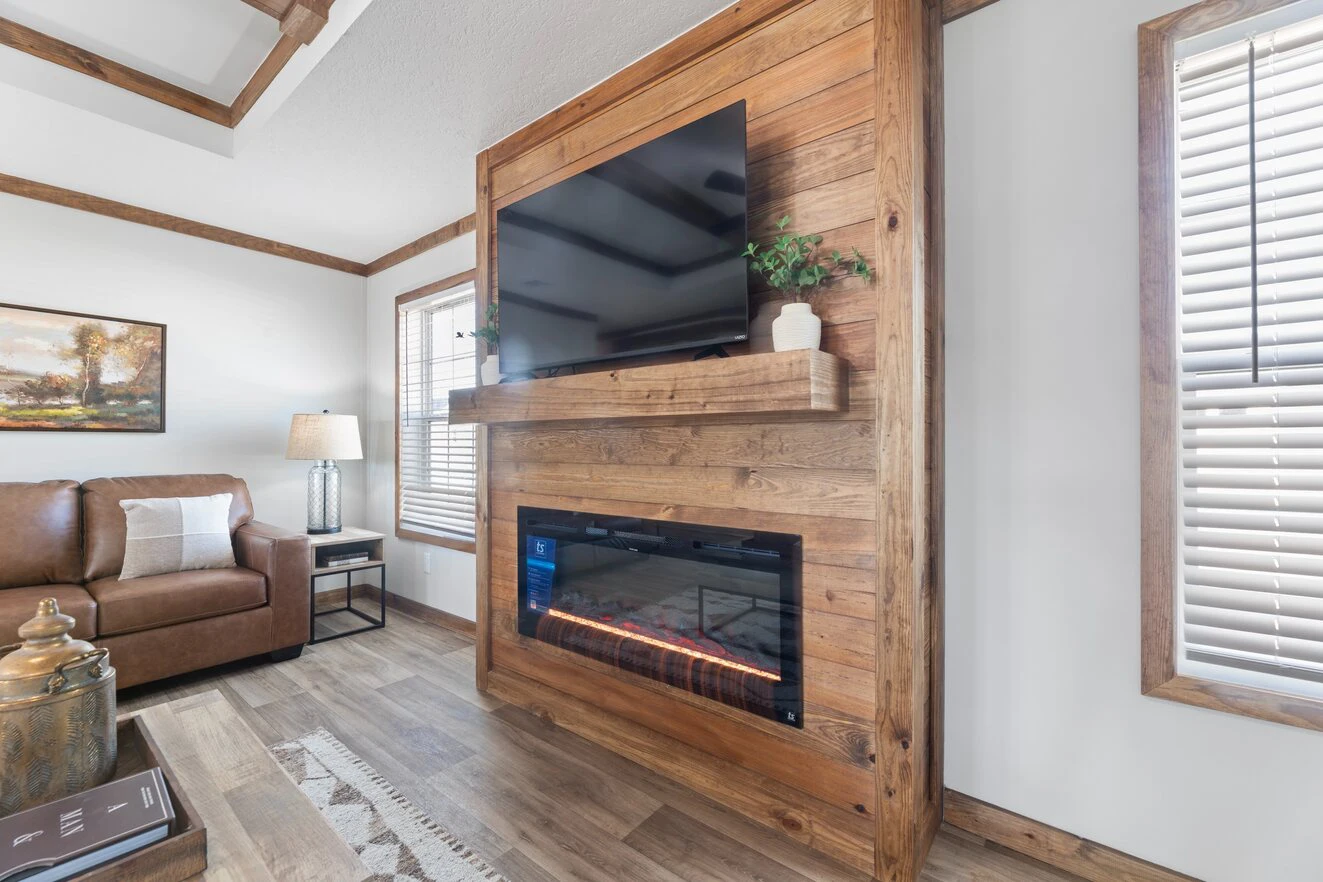 A living room features a wooden accent wall with a mounted TV and electric fireplace, a brown sofa, a side table with a lamp, and windows with blinds.