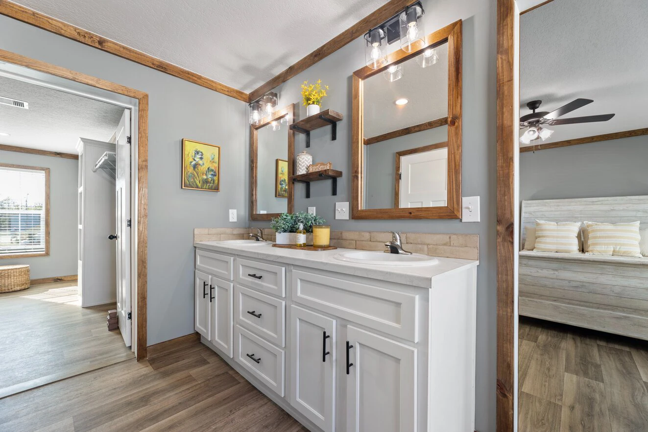 A bathroom with a double sink vanity, two mirrors, wall shelves, and wood trim, adjacent to a bedroom and hallway with wood flooring throughout.