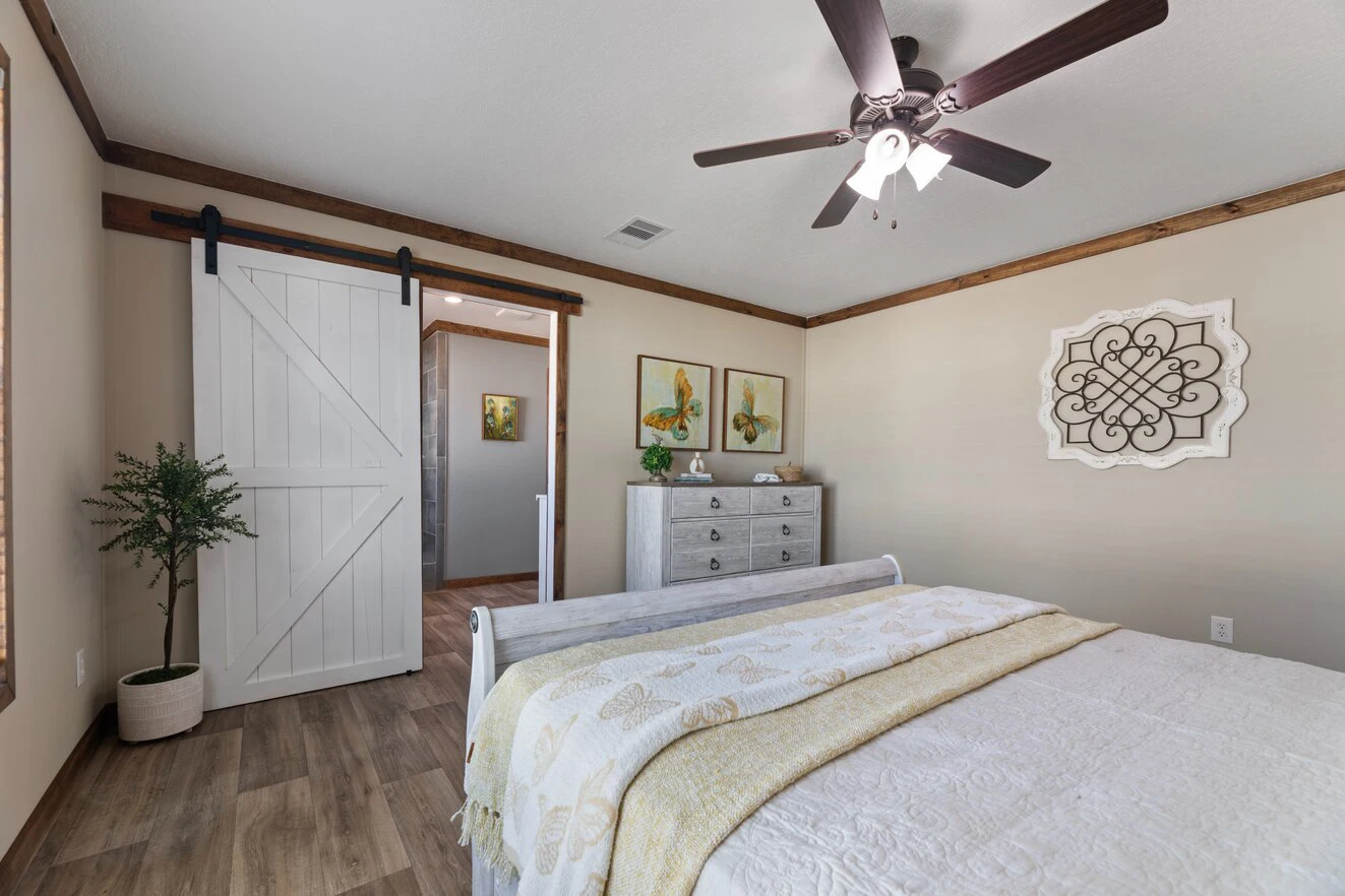 A bedroom with a white bed, light-colored bedding, a ceiling fan, a dresser with decor, a potted plant, and a white sliding barn door leading to another room.