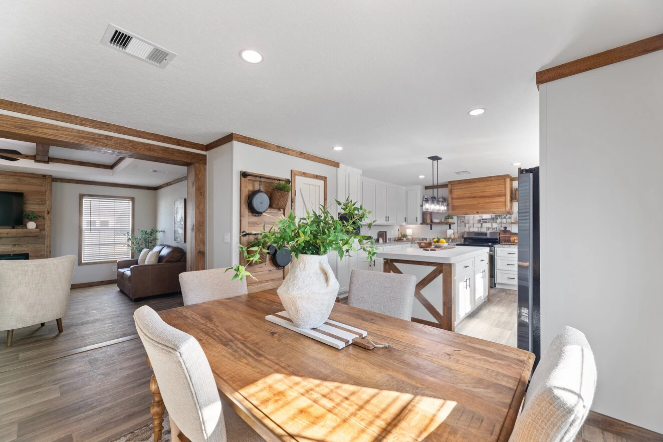 A bright open-plan kitchen and dining area with wooden accents, a vase of greenery on the table, and modern white cabinetry. The living room is visible in the background.
