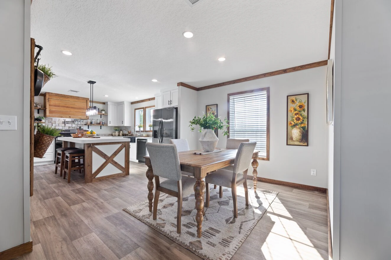 Bright kitchen and dining area with wood flooring, a wooden dining table with four chairs, and a kitchen island; sunlight streams through the window onto a patterned rug.