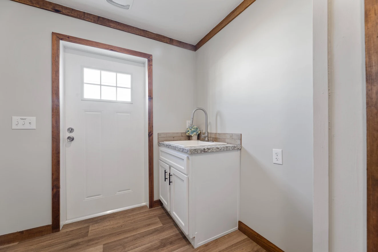 Small utility sink with cabinet and faucet next to a white exterior door in a room with light walls, wood trim, and wood flooring.