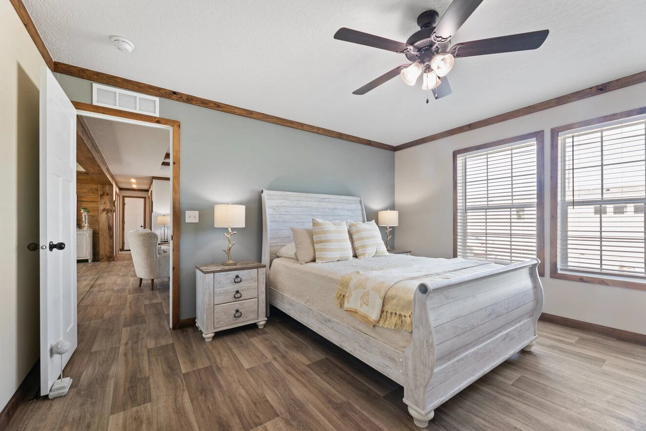 A bedroom with a white wooden bed, two matching nightstands with lamps, wood flooring, ceiling fan, and two large windows with blinds. Doorway opens to a hallway and living area.