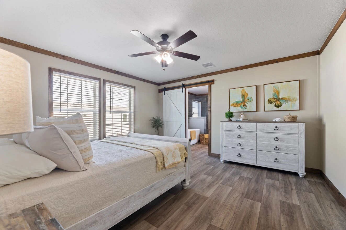 A bedroom with a wooden floor, bed, ceiling fan, dresser with two butterfly paintings above, and a sliding barn door leading to a bathroom.