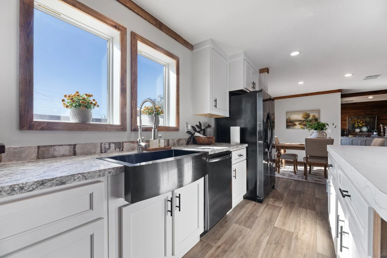 Modern kitchen with white cabinets, stainless steel farmhouse sink, and wood accents. Sunlight streams through two windows above the sink. Dining area visible in the background.