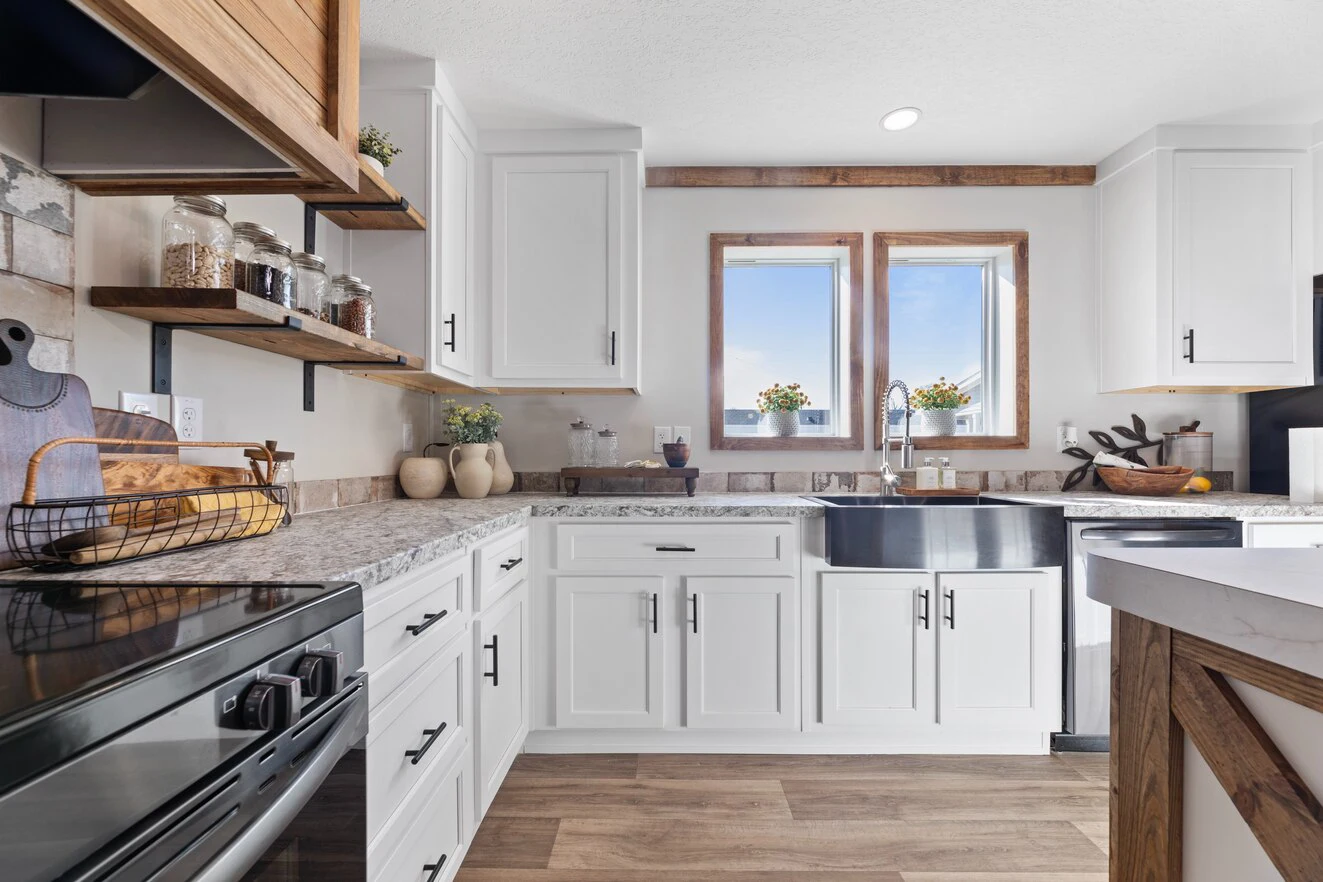 Modern kitchen with white cabinets, granite countertops, farmhouse sink, stainless steel appliances, and wooden accents; two windows above the sink provide natural light.