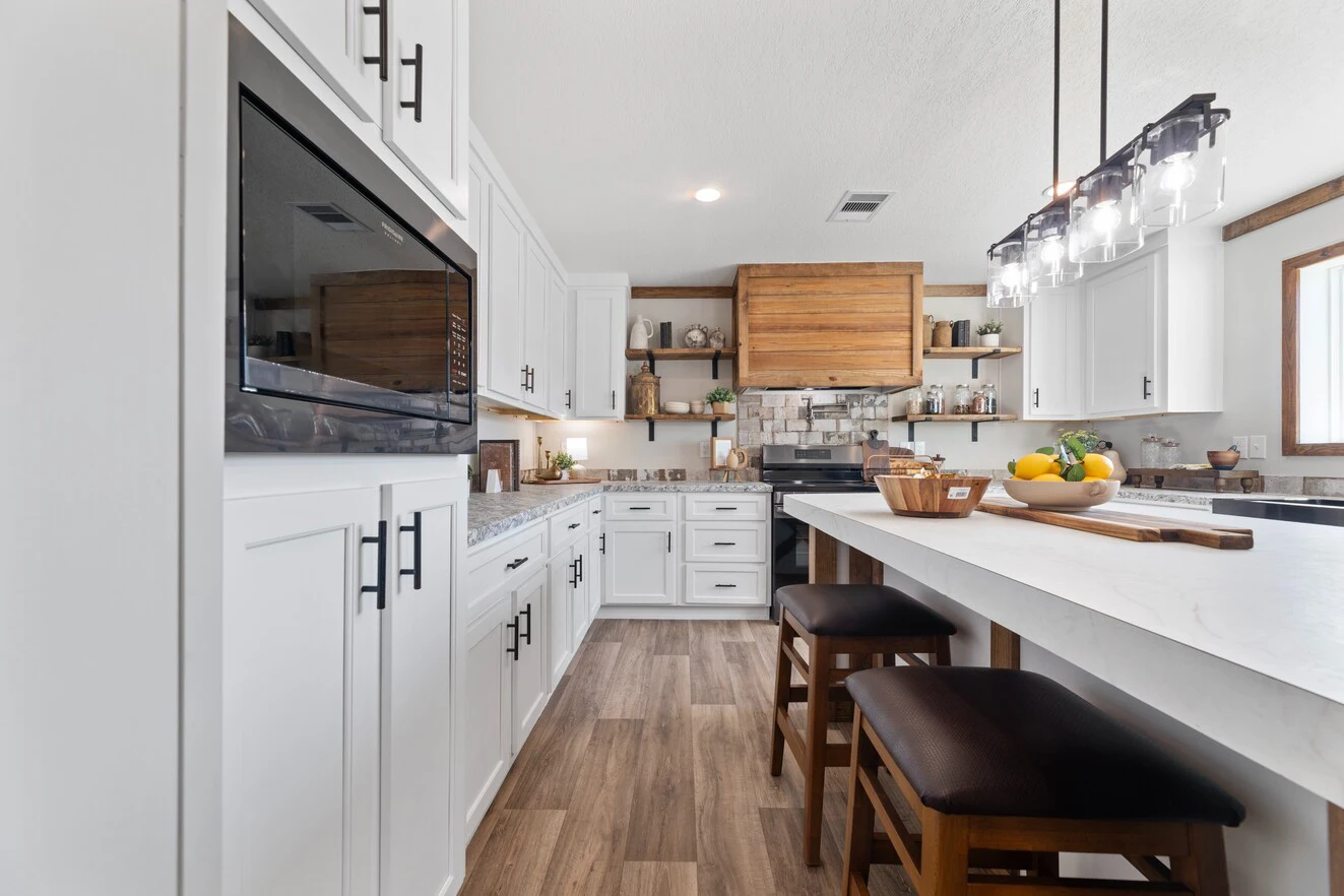 Modern kitchen with white cabinets, wood accents, a center island with two stools, pendant lighting, and stainless steel appliances. Bowl of fruit and kitchen items are on the countertop.