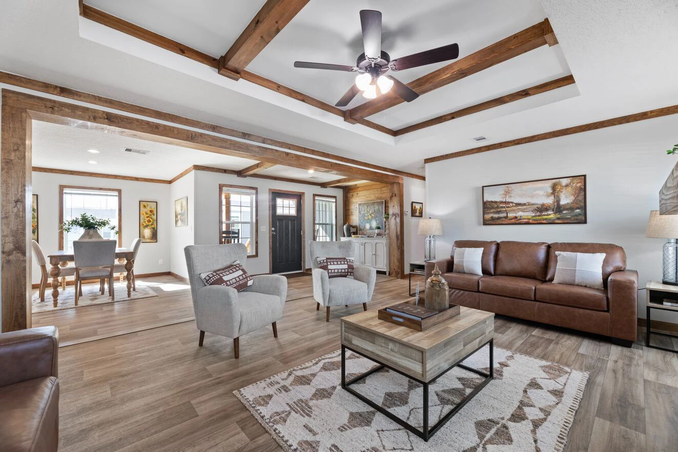Spacious living room with wood beams, ceiling fan, two armchairs, a brown leather sofa, coffee table, rug, and open view into dining area with large windows.
