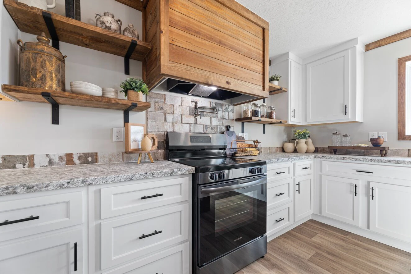 Modern kitchen with white cabinets, marble countertops, a black stove, wooden shelves, and a wooden range hood, featuring decorative jars and plants.