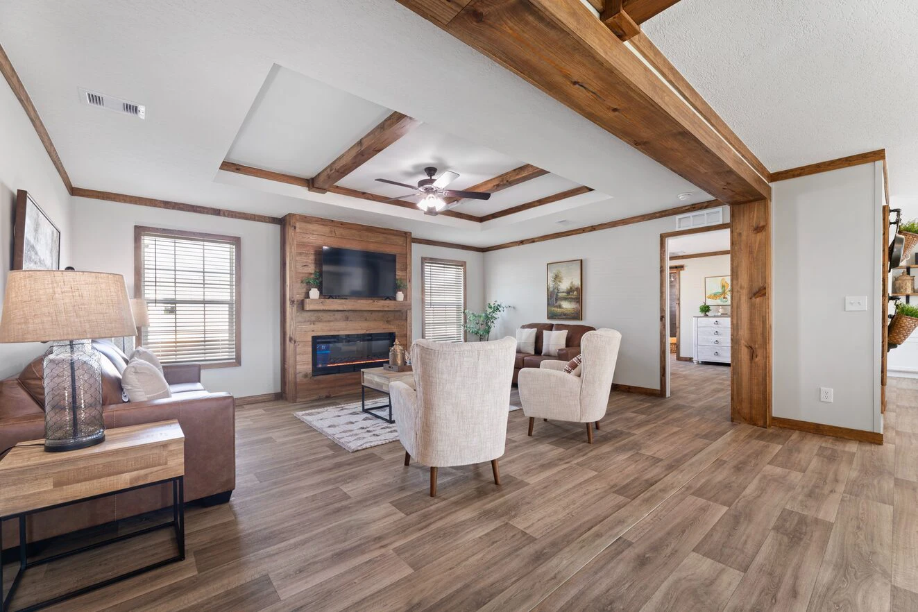 A living room with wood floors, exposed ceiling beams, a brown sofa, two beige armchairs, a wall-mounted TV above a fireplace, and large windows with wooden frames.