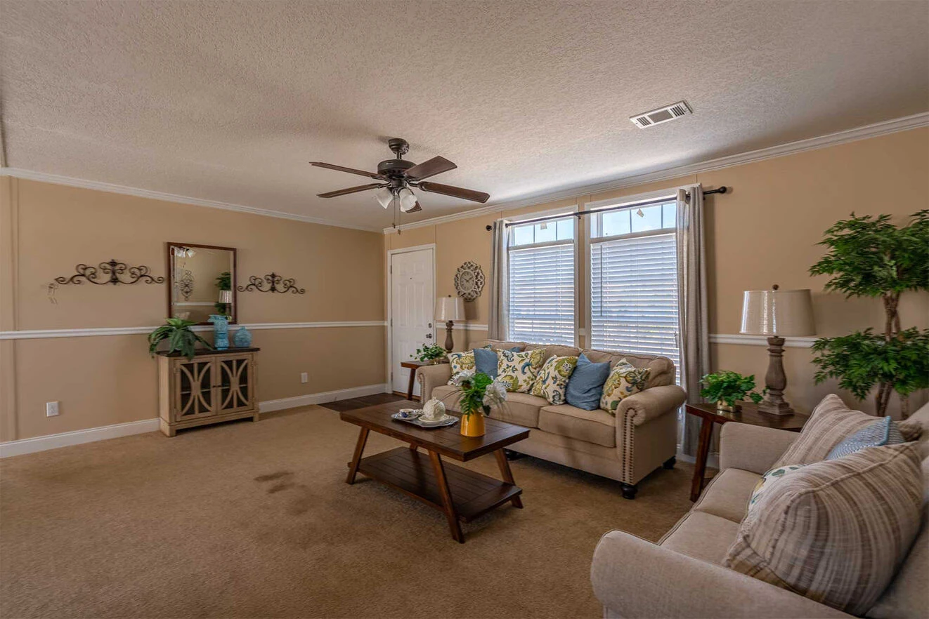te28523c_10_1713813643117_1323_12 A spacious living room with two beige sofas, a wooden coffee table, decorative plants, and large windows with blinds letting in natural light.