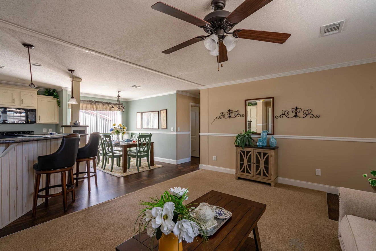 te28523c_11_1713813640427_1323_12 Open-plan living space with a kitchen, dining area, and living room. Features neutral tones, ceiling fan, bar stools, and decorative accents. Natural light from large windows.