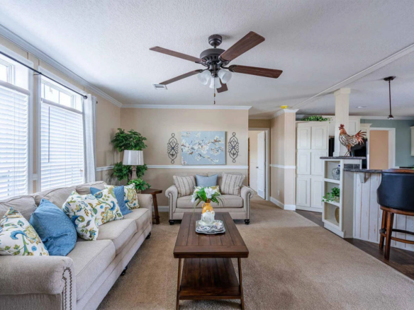 A living room with a sofa, armchairs, a wooden coffee table, decorative pillows, wall art, and a ceiling fan, adjacent to a kitchen with a breakfast bar and barstools.