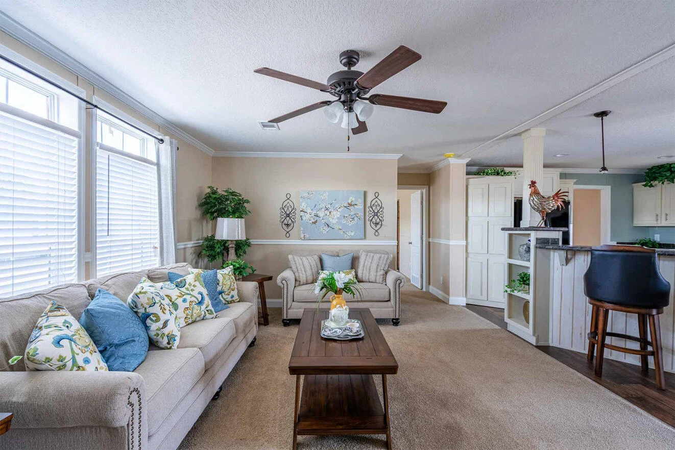 te28523c_12_1713813637388_1323_12 A living room with a sofa, armchairs, a wooden coffee table, decorative pillows, wall art, and a ceiling fan, adjacent to a kitchen with a breakfast bar and barstools.