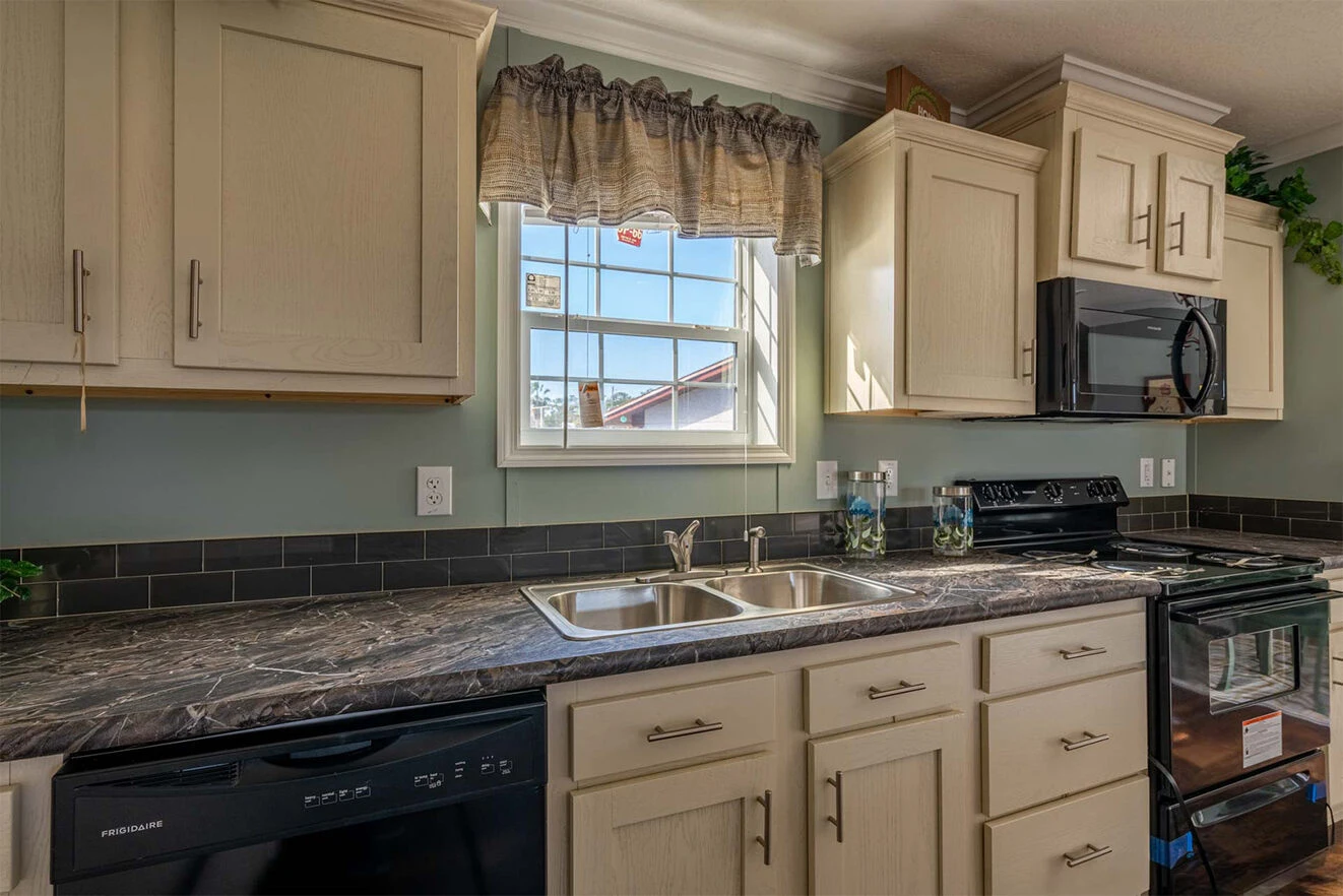 te28523c_13_1713813634063_1323_12 Modern kitchen with cream cabinets, dark countertops, a double sink under a window, black appliances including a stove and dishwasher, and a valance above the window.