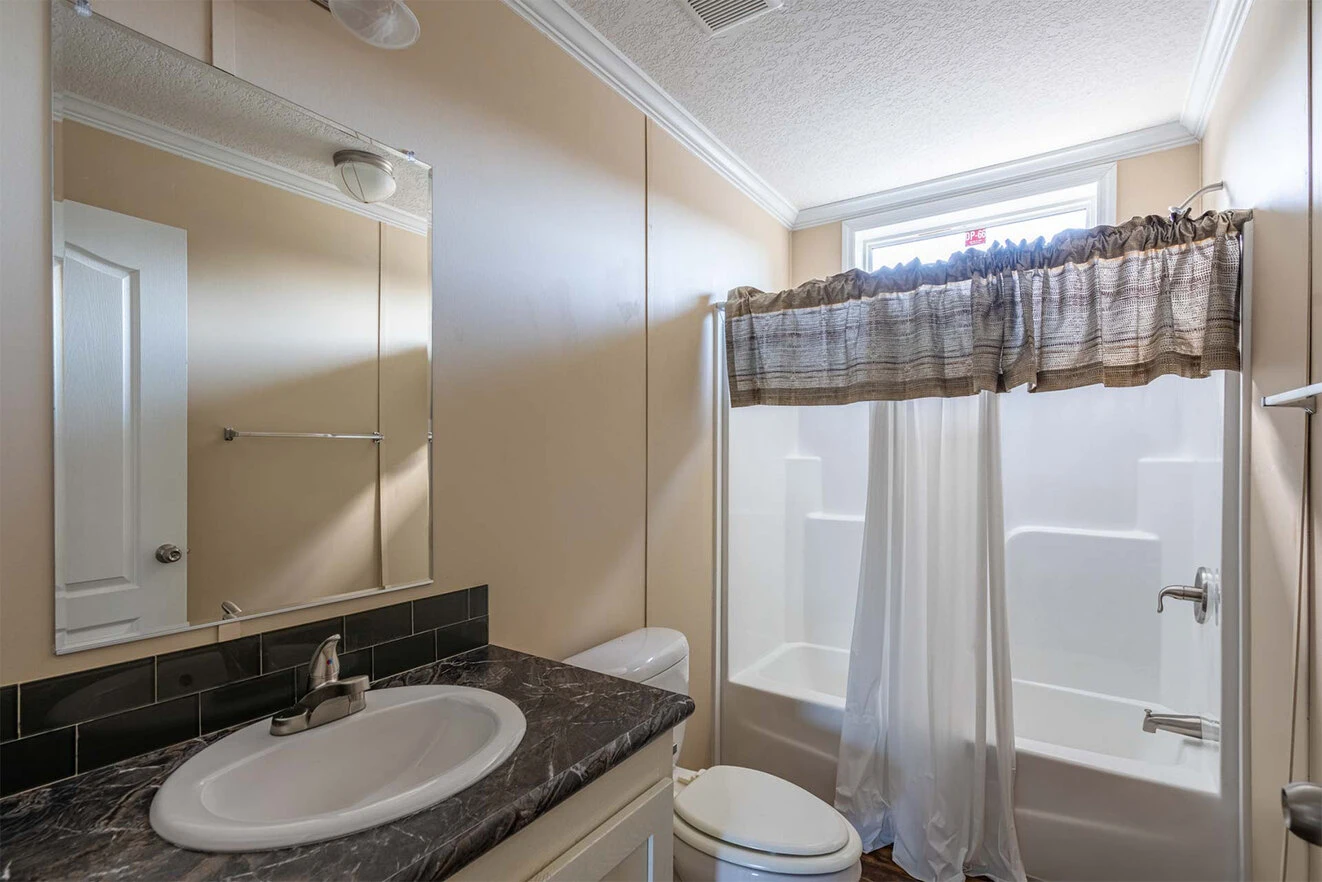 te28523c_4_1713813692518_1323_12 A small bathroom with a sink, toilet, and bathtub-shower combo, featuring beige walls, a dark countertop, and a window with a brown valance above the shower.