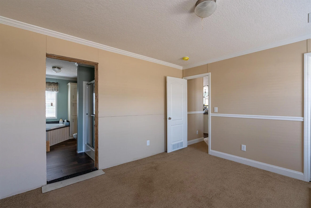 te28523c_5_1713813688826_1323_12 Empty room with beige walls, tan carpet, white trim, and two doorways—one open to a bathroom and another to a hallway. Ceiling light fixture visible.