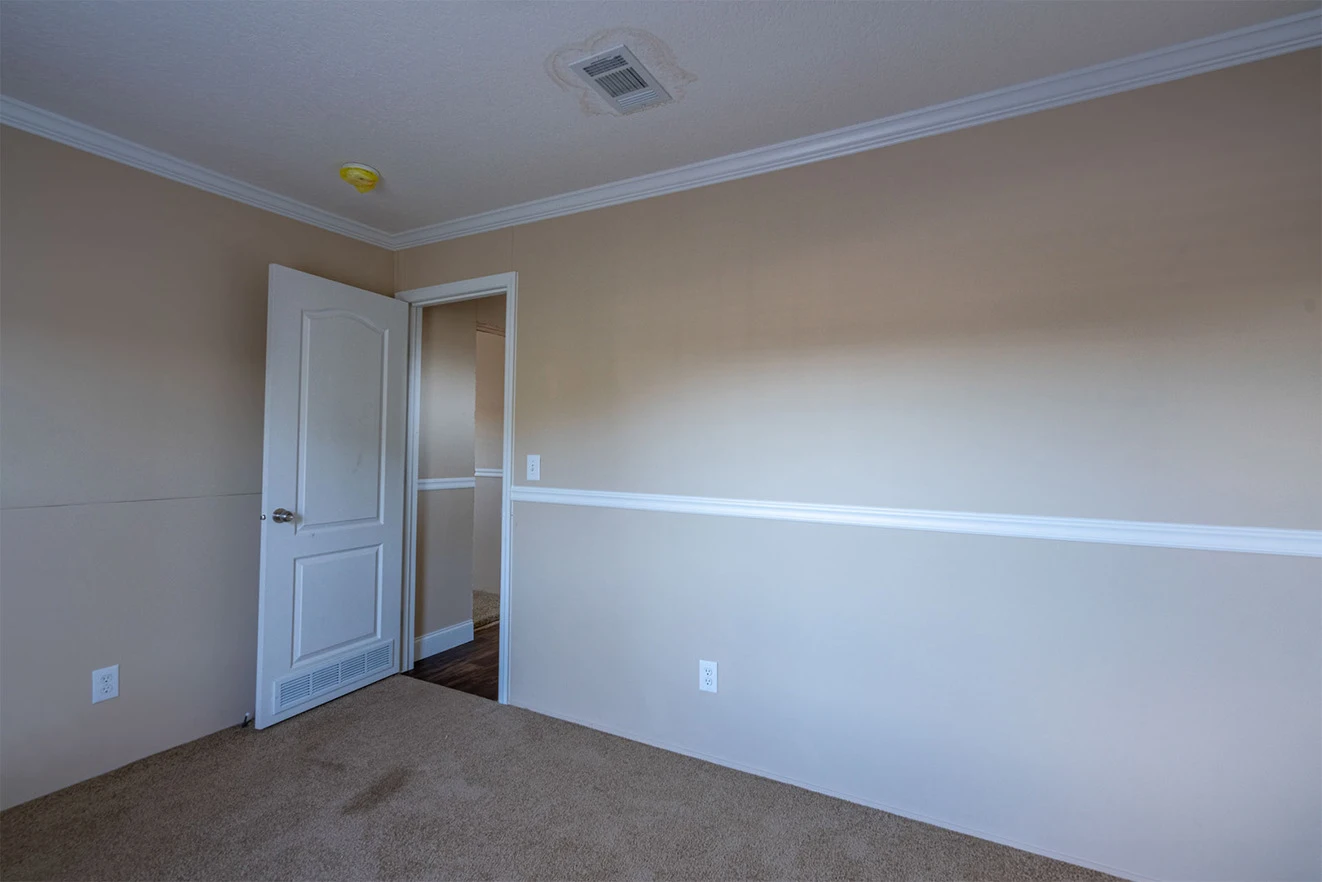 te28523c_7_1713813670204_1323_12 Empty room with beige walls, white trim, carpeted floor, and an open door leading to a hallway. Ceiling air vent and smoke detector are visible.