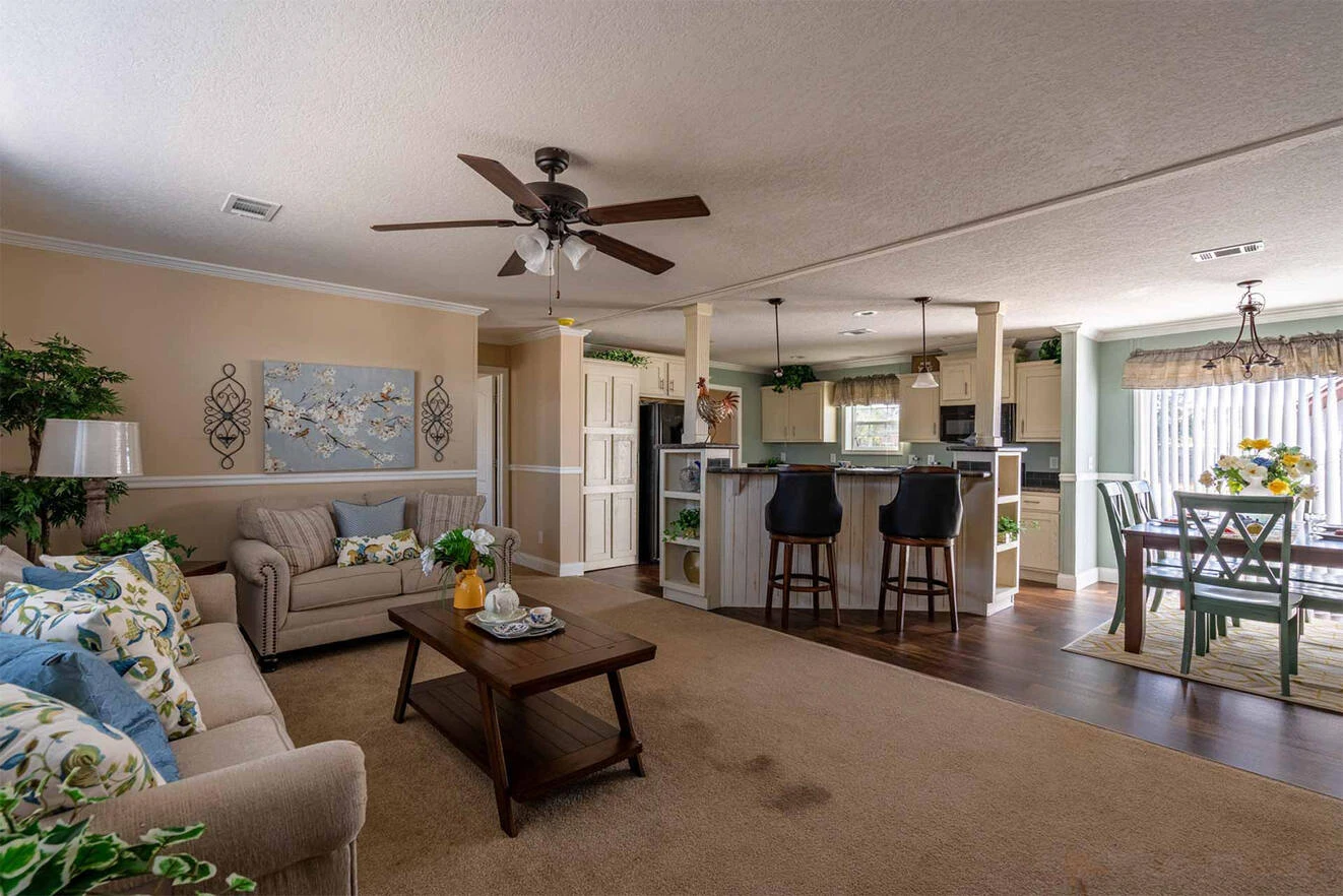 te28523c_8_1713813657193_1323_12 Open-plan living area with a beige sofa, coffee table, kitchen with bar stools, dining table by a window, ceiling fan, and decorative plants.
