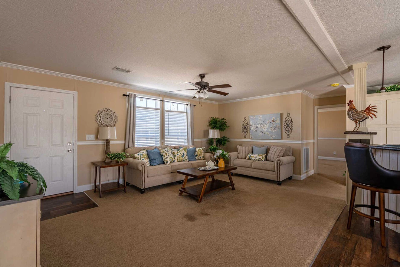 te28523c_9_1713813646211_1323_12 Spacious living room with beige walls, two sofas, a coffee table, ceiling fan, plants, and a bar area with stools in the background.