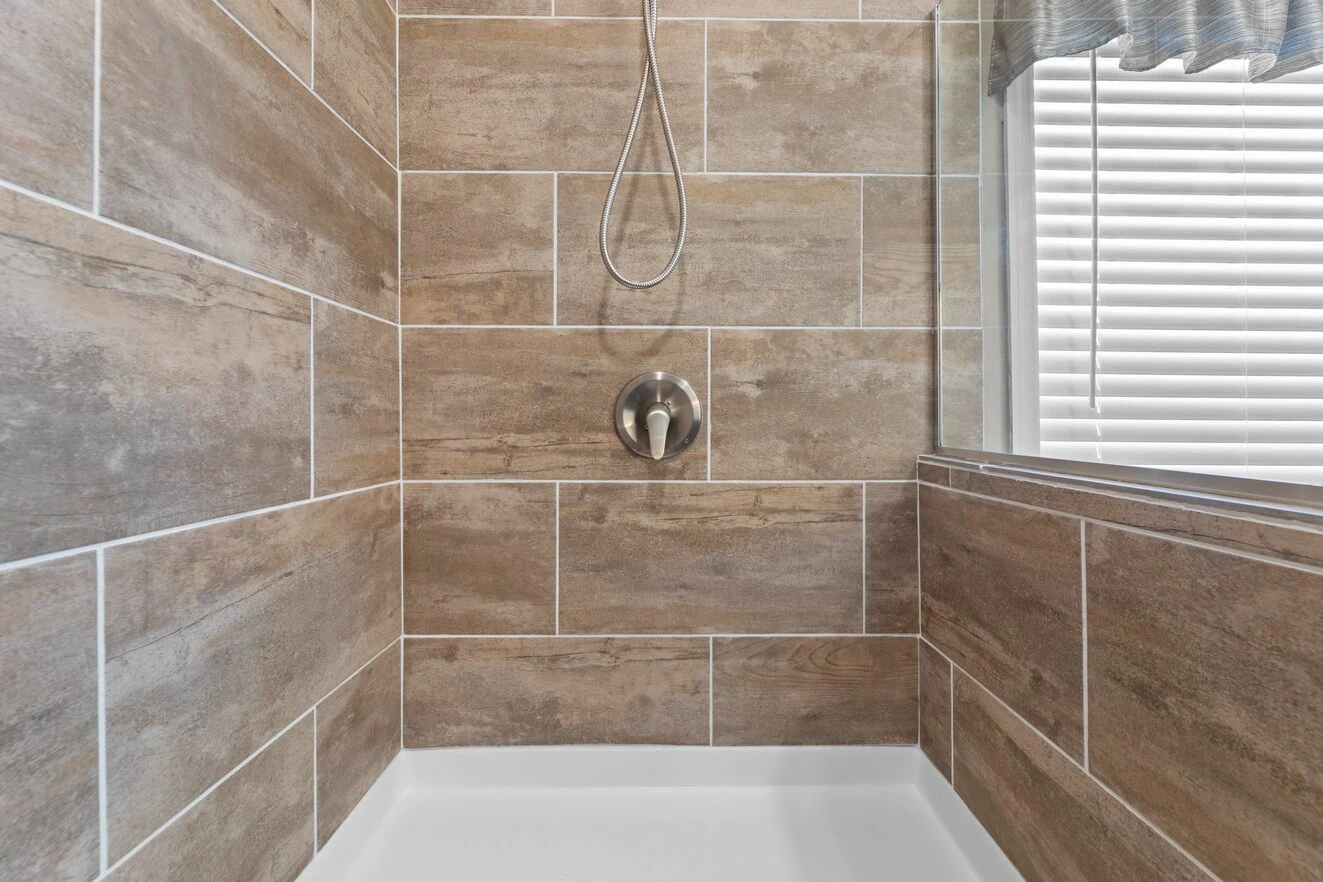 Shower area with tan and brown rectangular tiles, a silver shower handle, and a window with closed blinds on the right side.