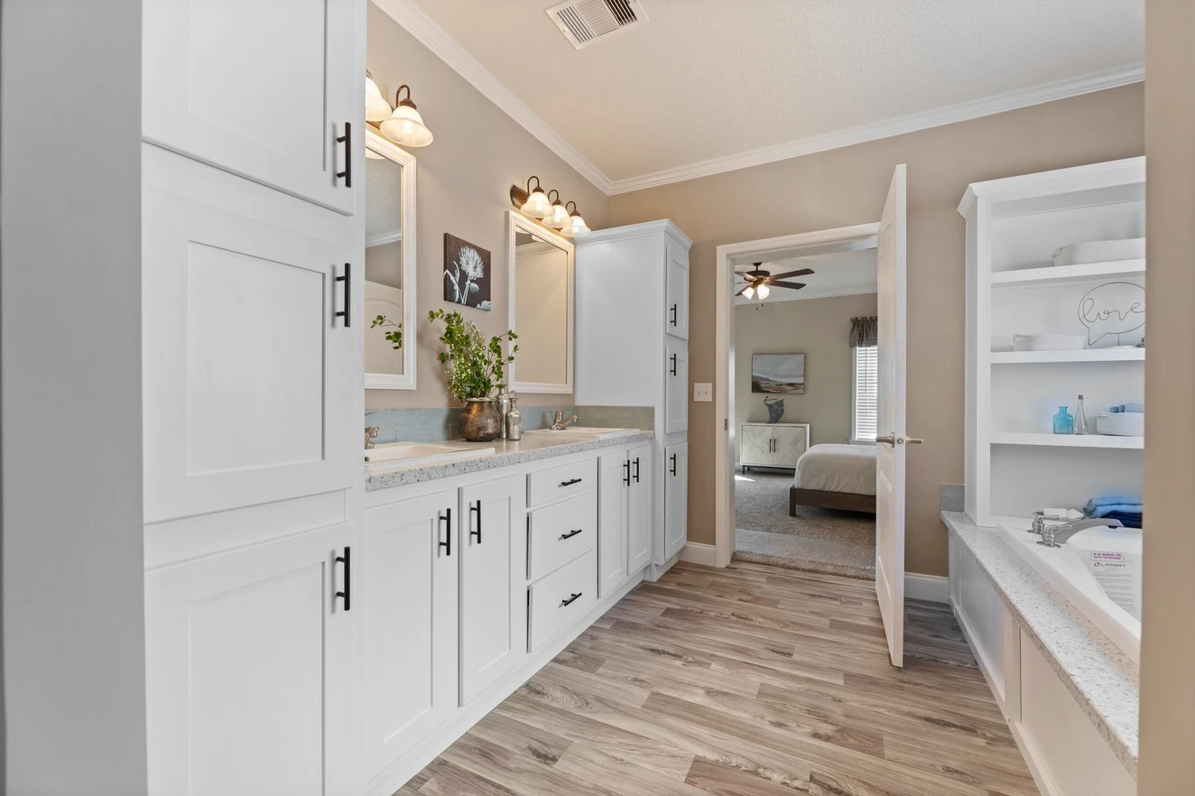 Modern bathroom with white cabinetry, double sinks, wall-mounted mirrors, wood-look flooring, built-in shelving, and an open door leading to a bedroom.