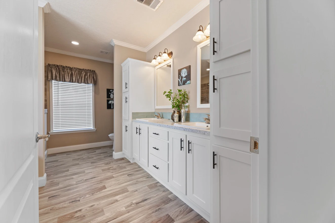 Bathroom with white cabinets, dual sinks, wall-mounted lights, a large mirror, wood-look flooring, and a window with a valance. A plant and framed art decorate the vanity.