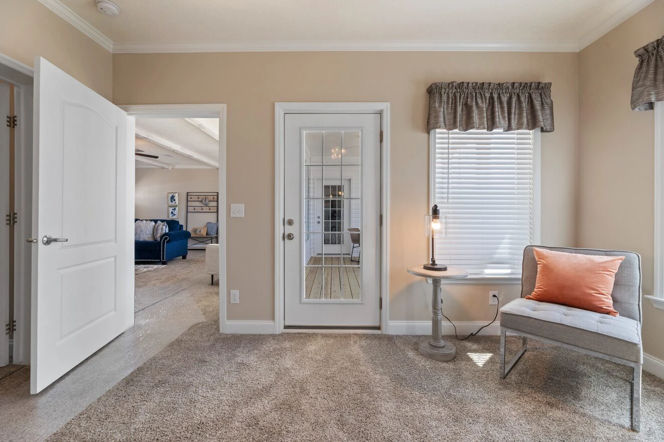A small room with beige walls and carpet, featuring a glass door, a window with blinds, a chair with an orange pillow, and a round table with a lamp.