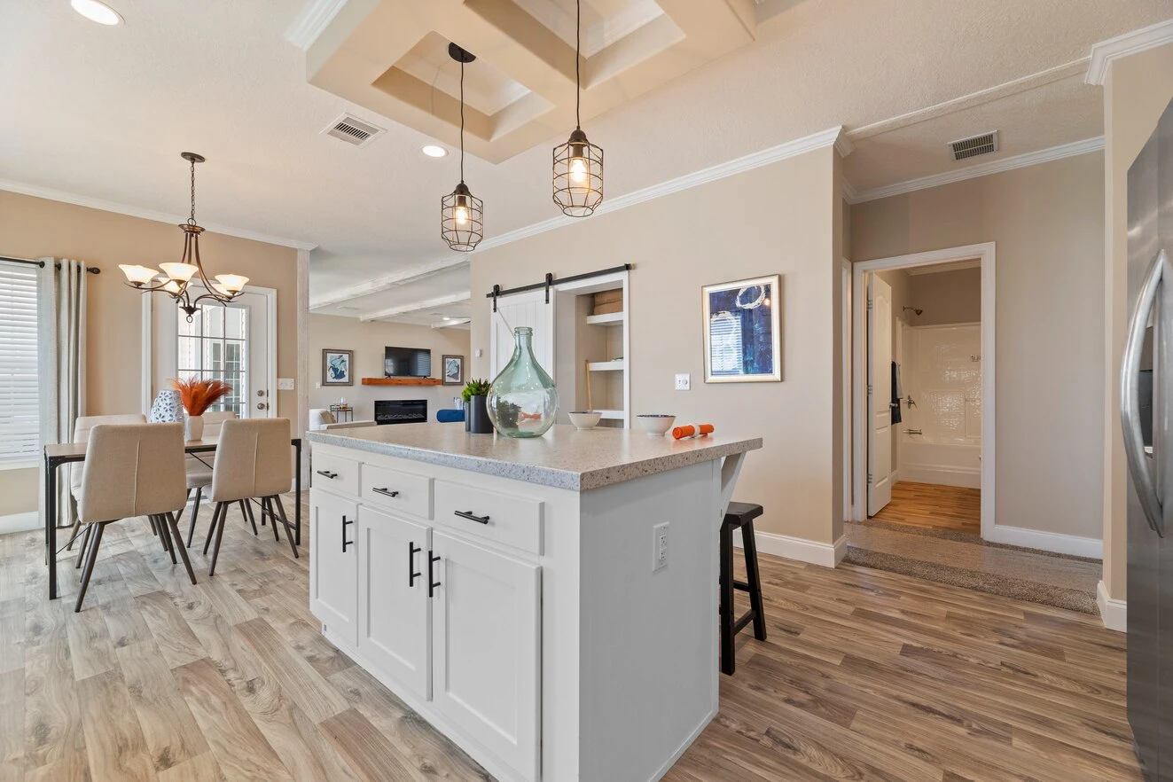 Modern kitchen and dining area with light wood flooring, white island, pendant lighting, and neutral-colored walls. Dining table and chairs are visible in the background.