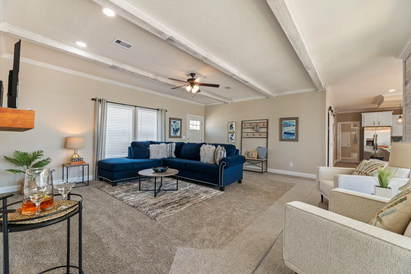 A spacious living room with a blue sectional sofa, two beige armchairs, a round coffee table, and neutral decor. A kitchen is visible in the background.