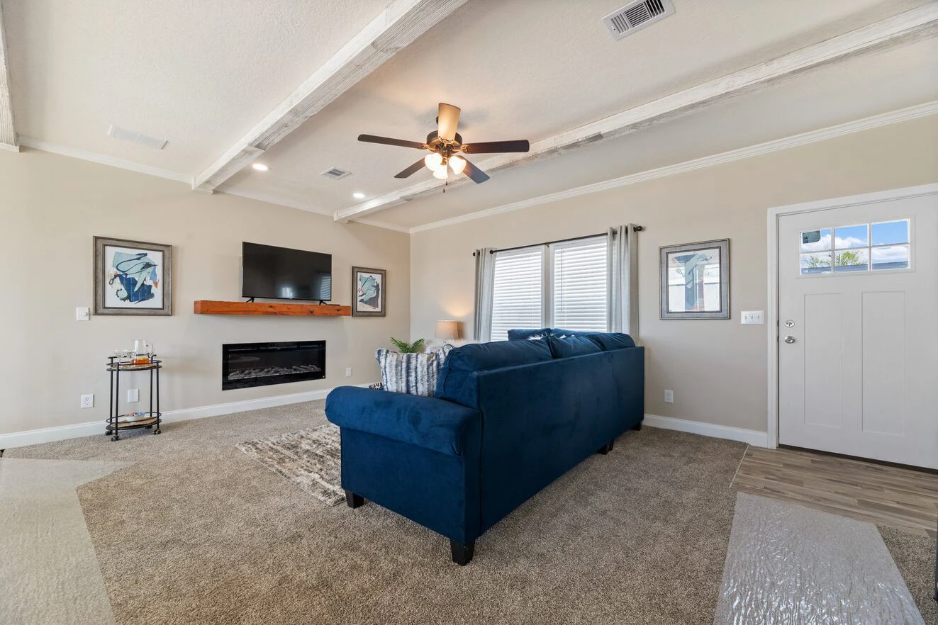 Living room with beige walls, ceiling beams, blue sofa, wall-mounted TV above a fireplace, large window with curtains, framed art, and a door leading outside.