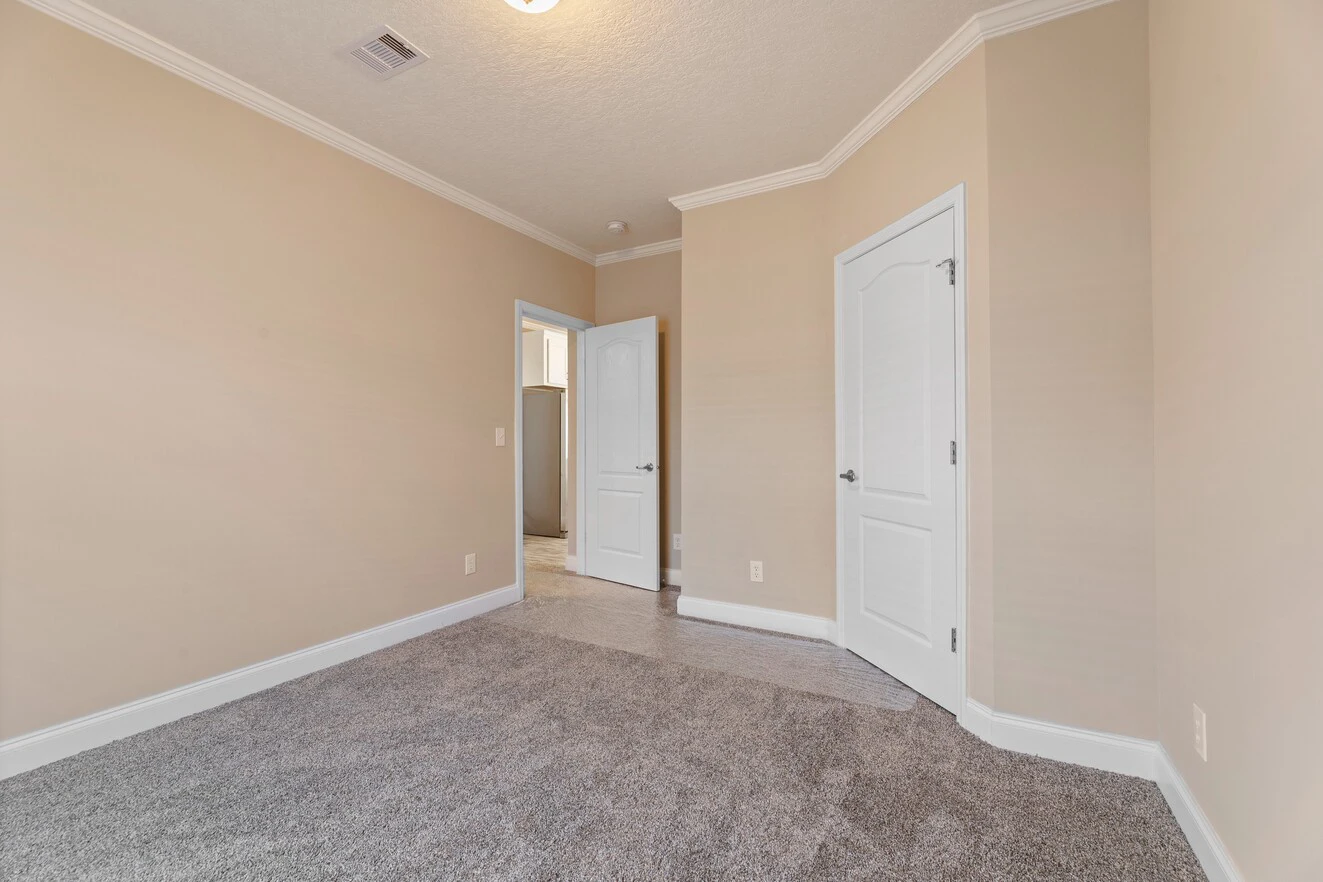 Empty beige-walled room with carpeted floor, white trim, two closed white doors, and one open door leading to another area. Ceiling light fixture is visible.