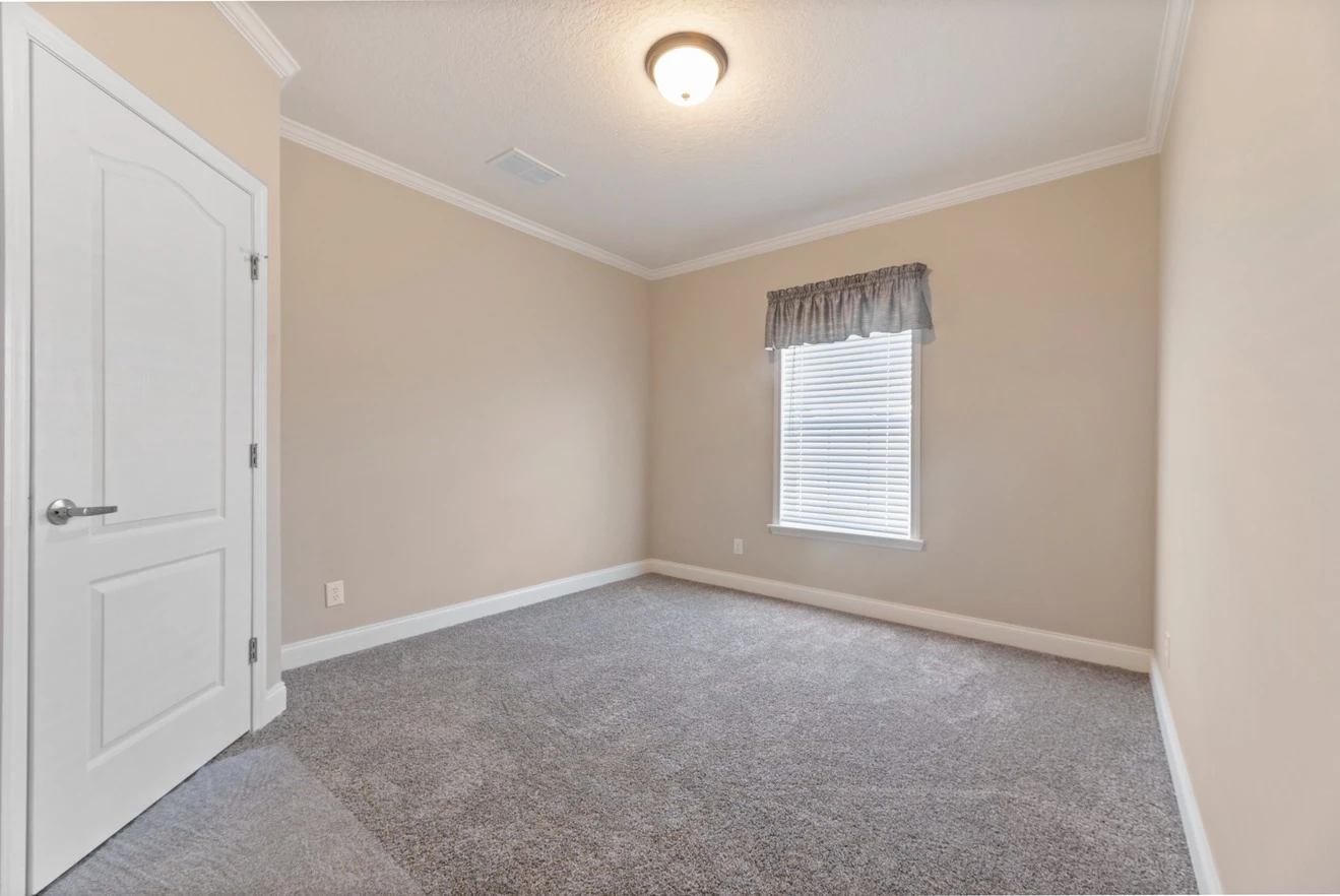Empty room with beige walls, gray carpet flooring, a single window with a valance, and a closed white door under a ceiling light fixture.