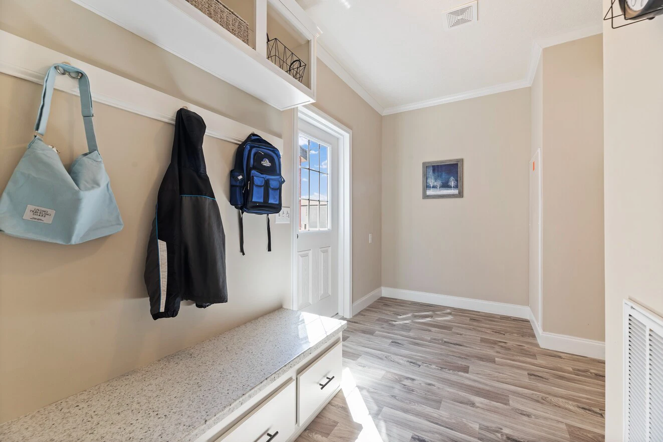 A mudroom with light wood flooring, beige walls, wall hooks holding two bags and a jacket, a cushioned bench with drawers, and a door leading outside.