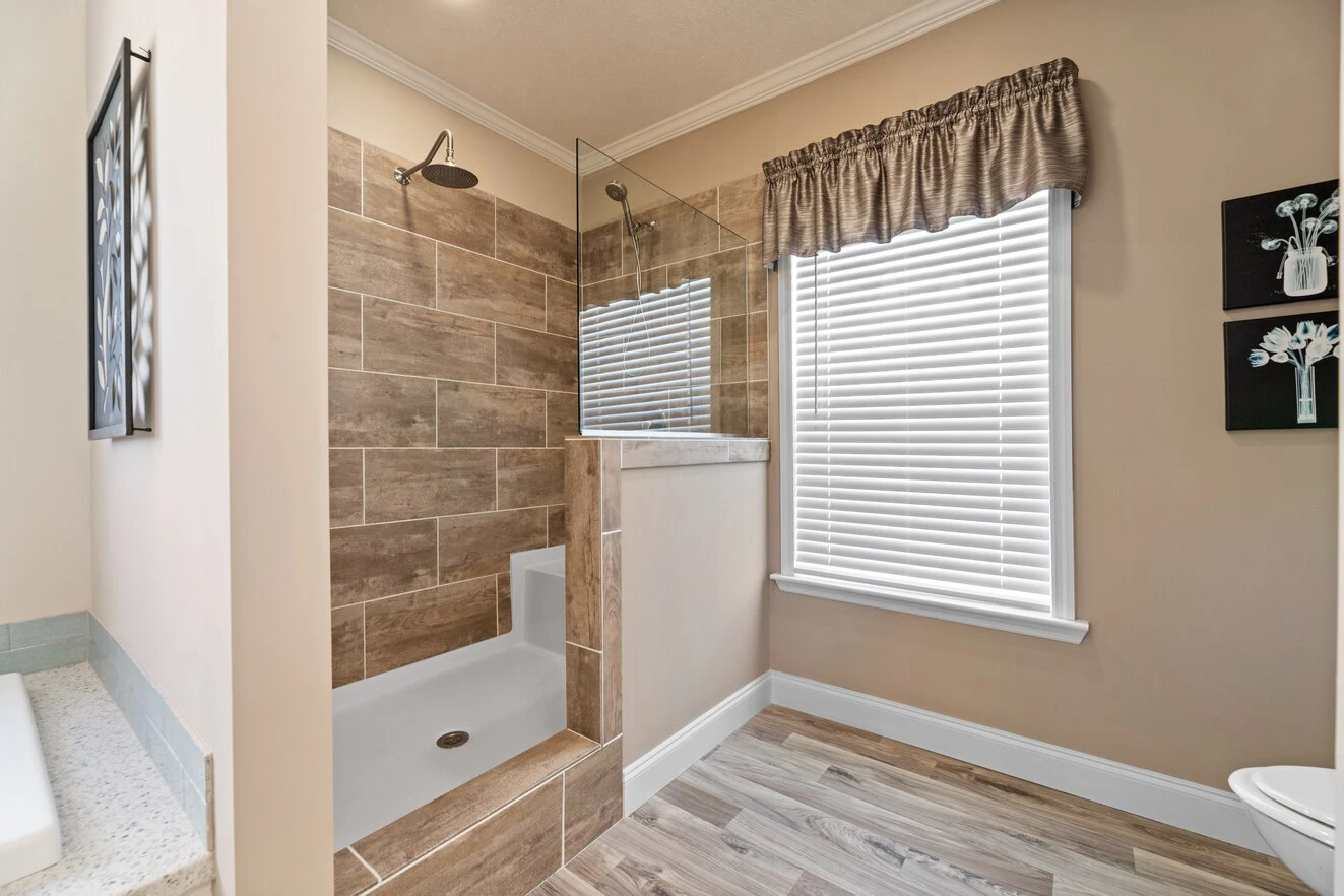 Walk-in shower with brown tiles and glass partition next to a window with blinds and valance in a bathroom with light wood flooring.