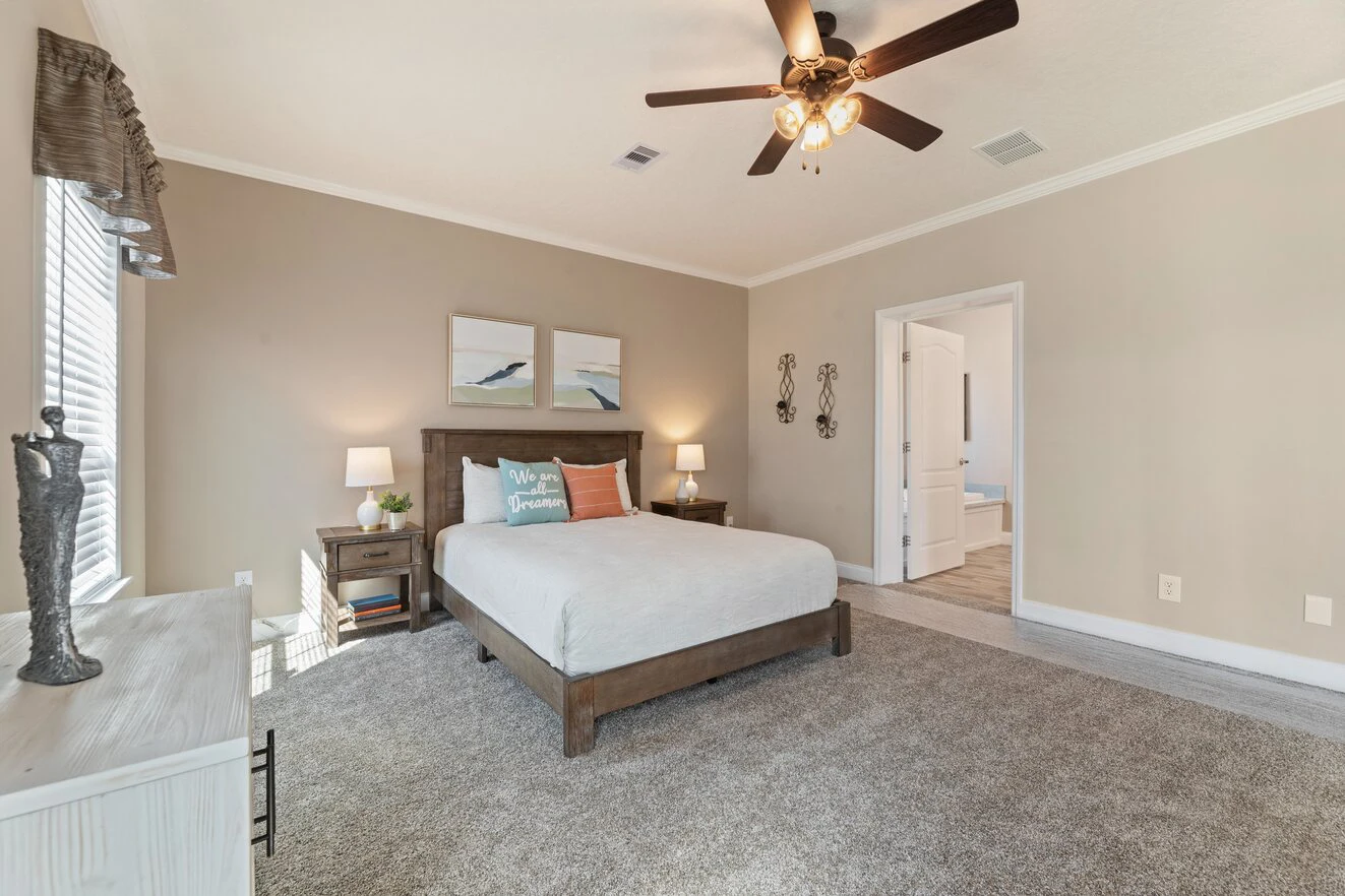 Spacious bedroom with a large bed, two nightstands, neutral walls, carpeted floor, ceiling fan, and wall art above the headboard. Doorway leads to an adjoining room.