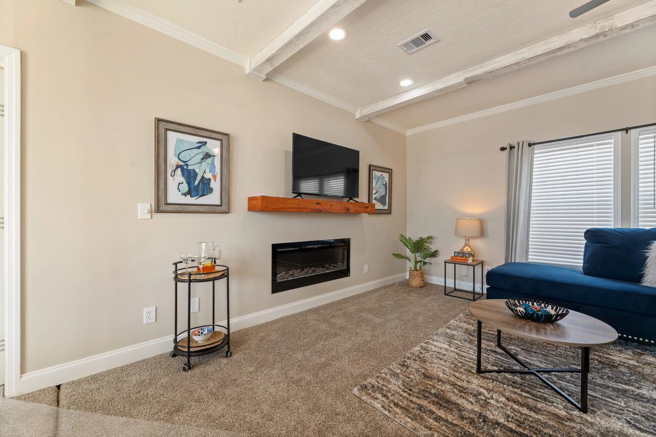 A living room with beige walls, a mounted TV above a fireplace, framed art, a blue sofa, a round coffee table, and a bar cart on carpeted flooring.