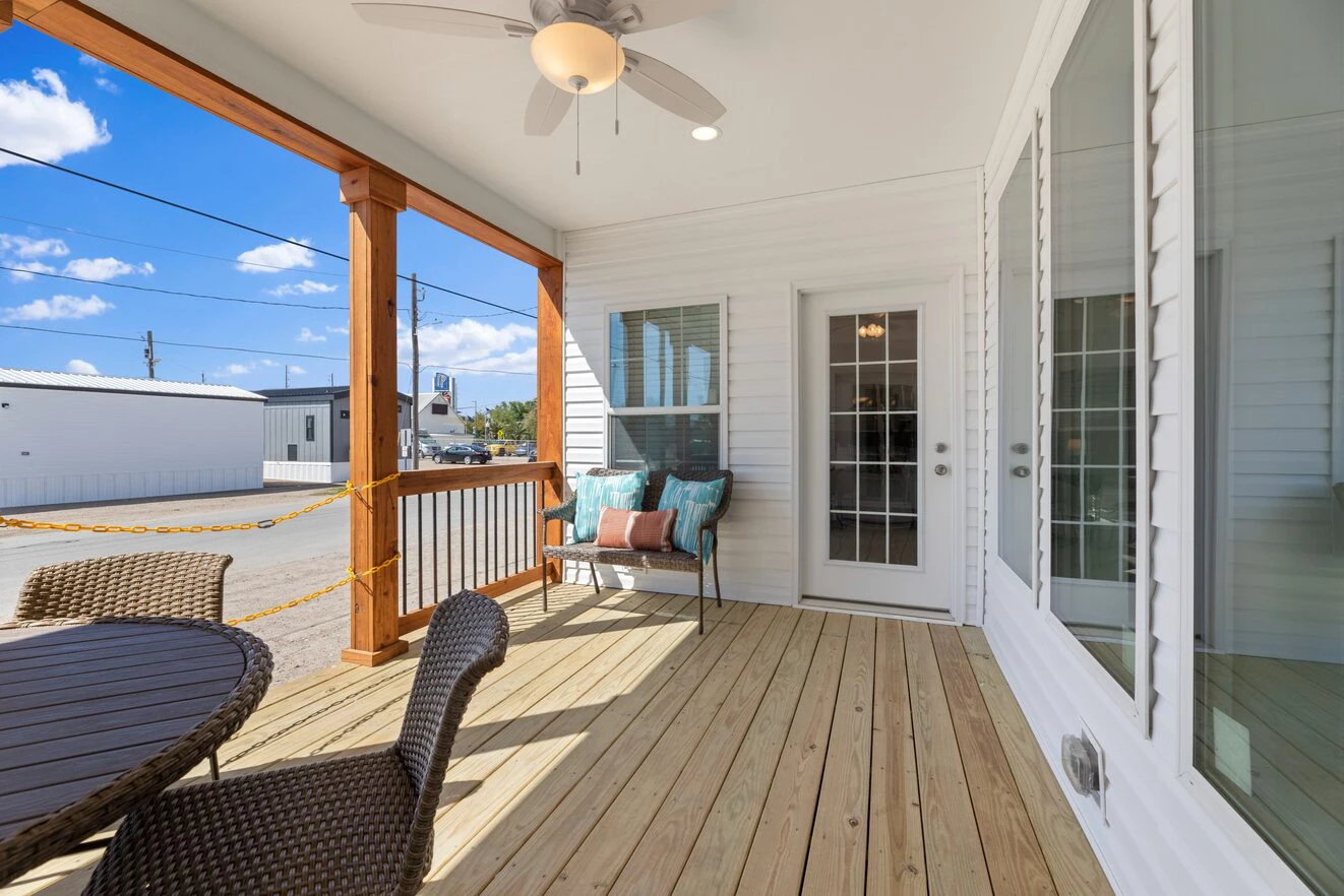 A wooden porch with a ceiling fan, wicker chairs around a table, and a cushioned bench, attached to a white house with glass doors and windows.