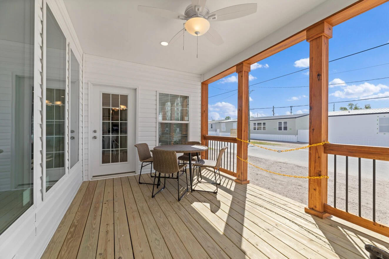 A covered wooden porch with a ceiling fan, table, and chairs, featuring wooden railings and a view of a street with mobile homes under a blue sky.